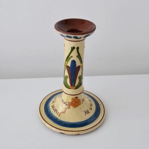 May include: A cream-coloured ceramic candlestick holder with a brown rim and base. The stem features a green, blue, and brown floral-inspired motif. The base is decorated with a blue circular band and the text, "He who has seven must..."