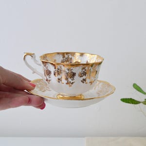 Pode incluir: Um conjunto de chávena e pires brancos com detalhes florais dourados. A borda e a alça da chávena são aparadas em ouro, assim como a borda do pires. A chávena é segurada em uma mão.