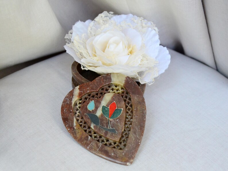 Puede incluir: Una caja de piedra de coraz&oacute;n, marr&oacute;n y blanca, con un dise&ntilde;o floral. La caja est&aacute; abierta y una flor de encaje blanco est&aacute; encima.