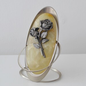 May include: A decorative oval-shaped object featuring a silver-colored metal frame and base. The center displays a light yellow stone with a silver rose design. The rose has two blooms and several leaves. The object is designed to stand upright.