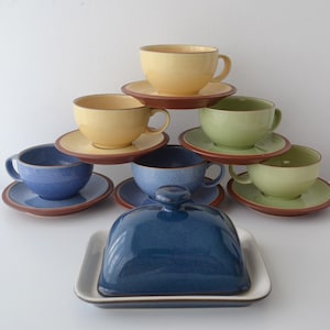 Op de afbeelding: Een verzameling keramische kopjes en schotels in tinten geel, groen en blauw, gerangschikt in een piramidevorm. Een blauwe boterpot met deksel staat aan de basis. De set heeft een rustieke, ambachtelijke uitstraling.