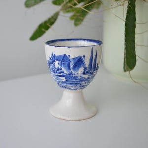 May include: A white ceramic egg cup with a blue and white landscape design. The design features houses, trees, and water. The egg cup has a pedestal base and a rounded top.