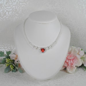 May include: A delicate pearl necklace with a red heart-shaped pendant. The pendant is surrounded by smaller rhinestones.