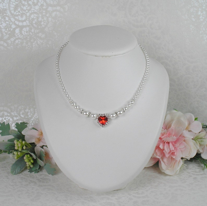 May include: A delicate pearl necklace with a red heart-shaped pendant. The pendant is surrounded by smaller rhinestones.
