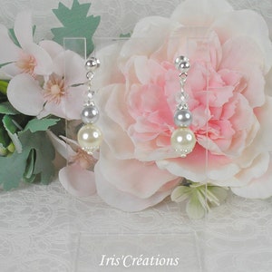 Peut inclure: Une paire de boucles d'oreilles pendantes en perles argentées et ivoire avec un accent de perle argentée et de cristal. Les boucles d'oreilles sont présentées sur un support en acrylique transparent sur un fond blanc avec des fleurs roses.