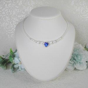 May include: A delicate silver necklace with a blue heart-shaped pendant surrounded by clear rhinestones. The necklace is made of small white pearls.