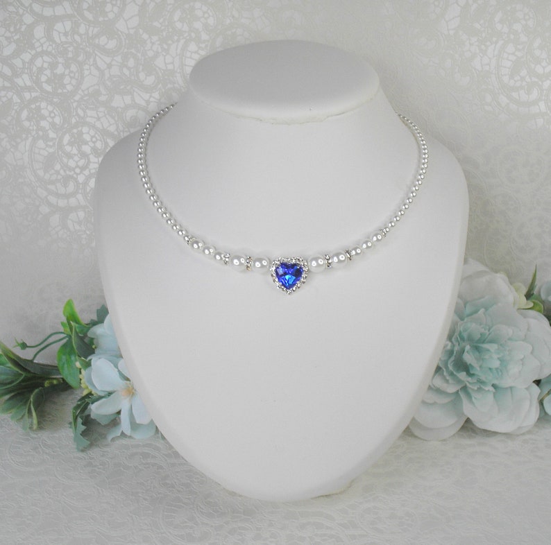 May include: A delicate silver necklace with a blue heart-shaped pendant surrounded by clear rhinestones. The necklace is made of small white pearls.