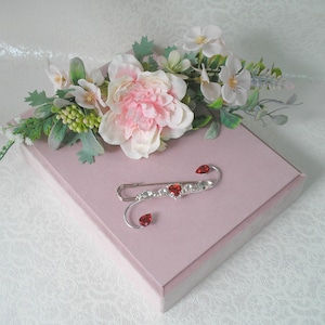 May include: A silver brooch, resembling a safety pin, is embellished with clear and red gemstones, set on a pink box. The brooch features heart-shaped and round stones. A floral arrangement with pink and white flowers sits on the box.