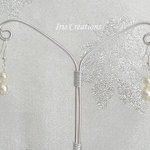 May include: A pair of dangle earrings with two off-white faux pearls each, suspended from silver-colored hooks. The earrings are displayed on a silver-colored jewelry stand against a shimmering, light-colored background. The text "Iris Creations" is visible.