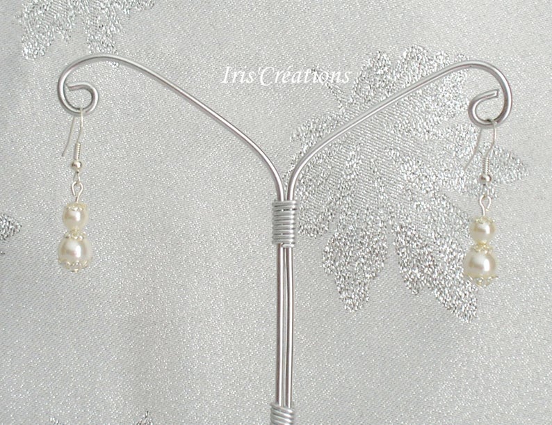 May include: A pair of dangle earrings with two off-white faux pearls each, suspended from silver-colored hooks. The earrings are displayed on a silver-colored jewelry stand against a shimmering, light-colored background. The text "Iris Creations" is visible.