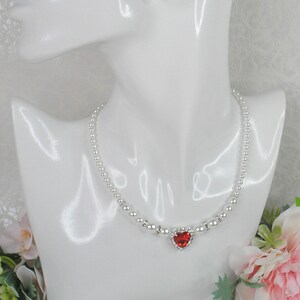 May include: A silver necklace with a heart-shaped pendant featuring a red gemstone. The necklace is made of small, round beads.