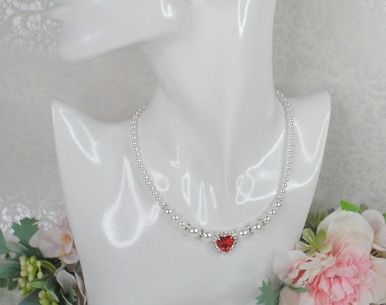 May include: A silver necklace with a heart-shaped pendant featuring a red gemstone. The necklace is made of small, round beads.