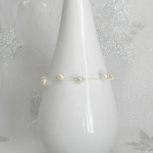 May include: A white ceramic vase with a slender, conical shape. A delicate bracelet with several cream-colored pearls is wrapped around the vase. The background is a soft, textured white with subtle silver floral accents. The text "Iris' Cr&eacute;ations" is visible at the bottom.