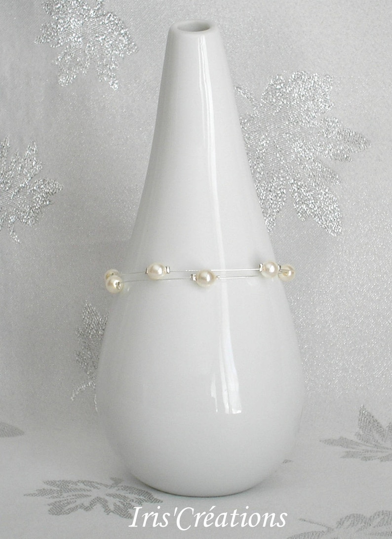 May include: A white ceramic vase with a slender, conical shape. A delicate bracelet with several cream-colored pearls is wrapped around the vase. The background is a soft, textured white with subtle silver floral accents. The text "Iris' Cr&eacute;ations" is visible at the bottom.