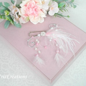 May include: A delicate white feather and pearl brooch with a pink ribbon and silver safety pin. The brooch is displayed on a pink velvet box.