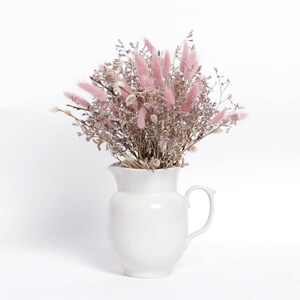 May include: A white ceramic pitcher filled with a bouquet of dried pink and white flowers. The flowers are arranged in a loose, natural style, and the pitcher has a simple, elegant design.