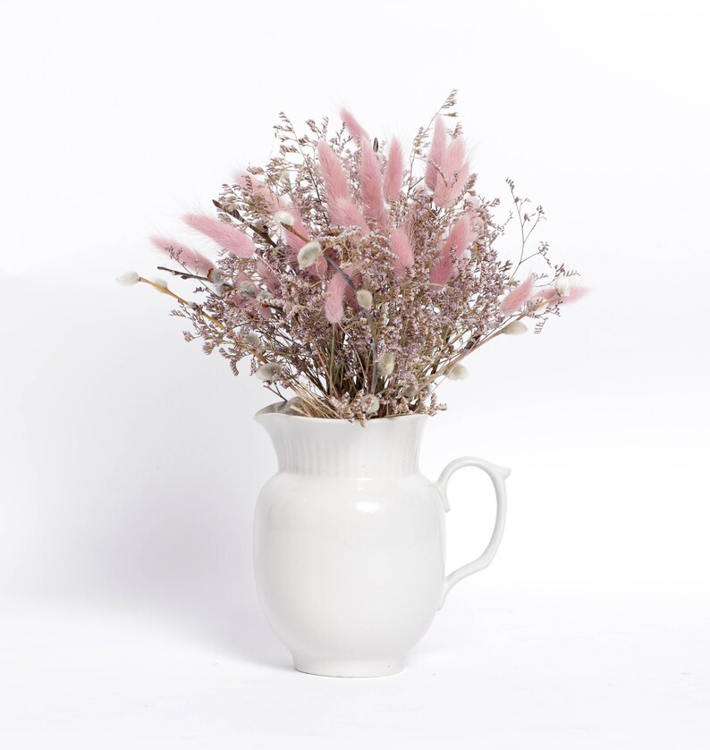May include: A white ceramic pitcher filled with a bouquet of dried pink and white flowers. The flowers are arranged in a loose, natural style, and the pitcher has a simple, elegant design.