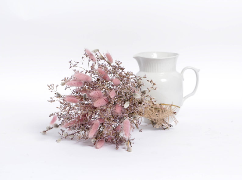 May include: A bouquet of dried pink bunny tail grass and white pussy willow branches tied with twine, next to a white ceramic pitcher.