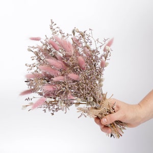 May include: A bouquet of dried pink and white flowers, tied with twine. The bouquet is held in a hand.