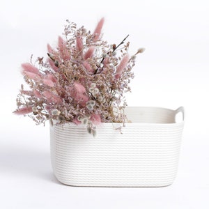 May include: A white rectangular basket with rope-like texture and two handles, filled with a bouquet of dried pink and white flowers.