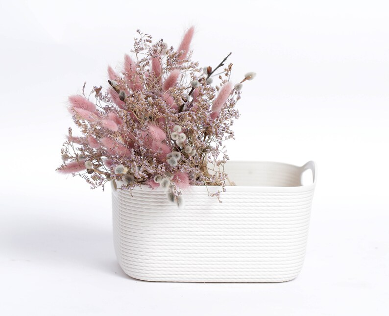 May include: A white rectangular basket with rope-like texture and two handles, filled with a bouquet of dried pink and white flowers.