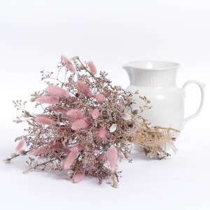May include: A bouquet of dried pink bunny tail grass and other dried flowers arranged in a white ceramic pitcher.