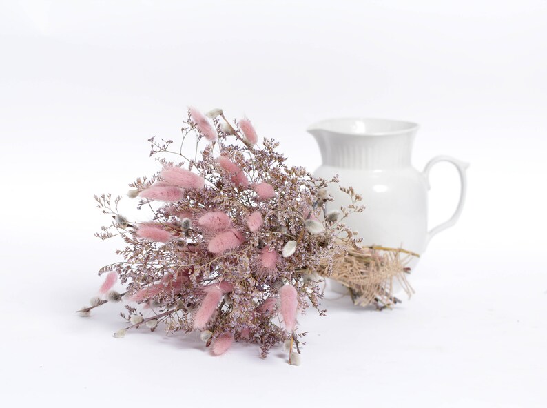 May include: A bouquet of dried pink bunny tail grass and other dried flowers arranged in a white ceramic pitcher.