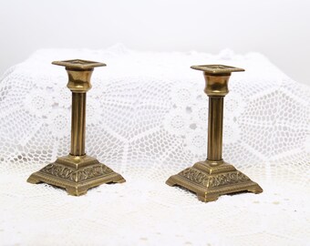May include: Two antique brass candle holders with ornate details on the base and a square top. The candle holders are on a white lace tablecloth.