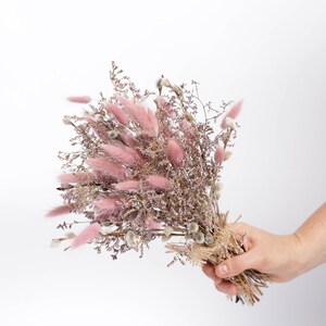 May include: A bouquet of dried pink and purple flowers tied with twine. The bouquet is held in a hand.