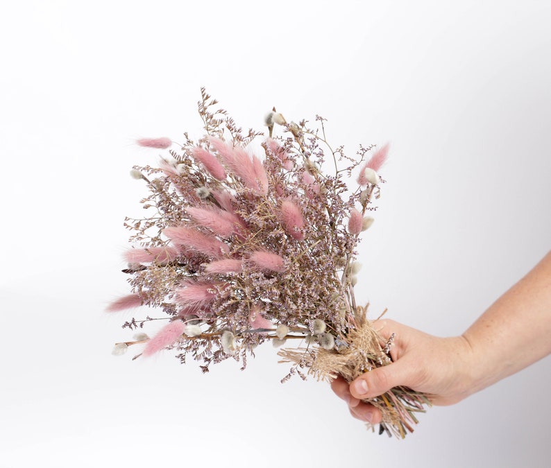 May include: A bouquet of dried pink and purple flowers tied with twine. The bouquet is held in a hand.