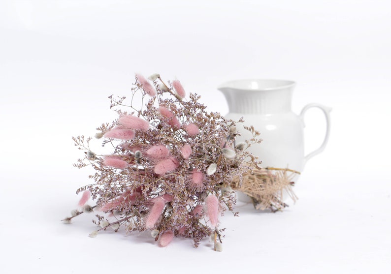 May include: A bouquet of dried pink bunny tail grass and dried willow branches arranged in a white ceramic pitcher.