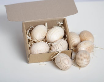 May include: A collection of wooden Easter eggs, some inside a cardboard box filled with straw, and others scattered around. The eggs have a natural wood grain pattern and are light brown in color.