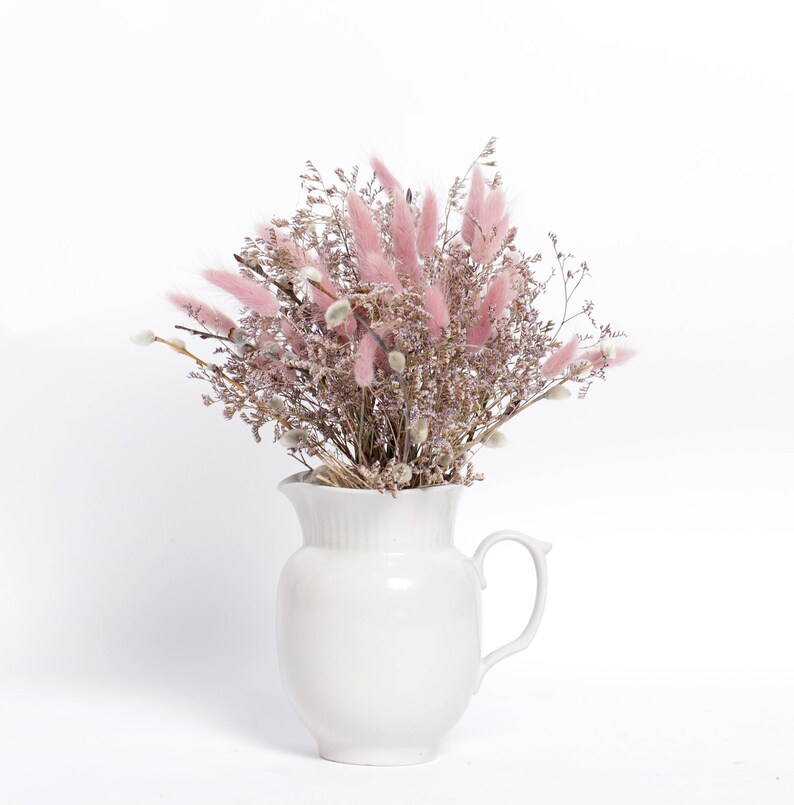 May include: A white ceramic pitcher filled with a bouquet of dried pink and white flowers. The flowers are arranged in a vase with a white background.