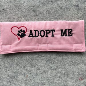 May include: Pink rectangular fabric with the embroidered text "ADOPT ME". A red heart and black paw print are embroidered on the left side. The fabric has stitched lines and tabs on the ends.