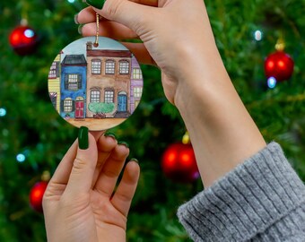 Può includere: Un ornamento natalizio rotondo in ceramica con un dipinto ad acquerello di case a schiera colorate. L'ornamento ha un cordino dorato per appenderlo e viene tenuto contro un albero di Natale con ornamenti rossi.