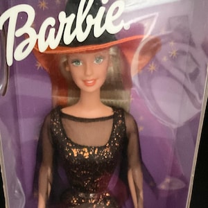 May include: A Barbie doll in a clear plastic box. The doll is dressed as a witch, wearing a black dress with gold sequins, a black hat with an orange brim, and holding a pink bag with a black cat design. The box has the word "Barbie" on top.
