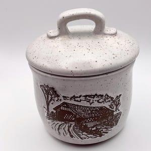 May include: A ceramic canister with a lid featuring a brown embossed graphic of a covered bridge. The canister has a light cream-colored base with small brown speckles throughout the surface and a handle on top of the lid.
