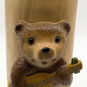 Puede incluir: Un recipiente de madera con un oso tallado tocando el ukelele. El oso es marrón con ojos negros y está adherido al recipiente marrón claro. La parte superior del recipiente es de color marrón oscuro.