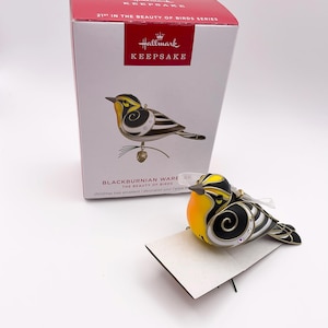 May include: A Hallmark Keepsake Blackburnian Warbler Christmas tree ornament featuring a black, white, and yellow bird with a small pinecone accent. The ornament is displayed in front of its original white and red box labelled 21st in the Beauty of Birds series.