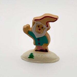 May include: A small, wooden gnome figurine with a tan hat and beard, a teal jacket, and pink pants. The gnome is waving and stands on a small, off-white base with a green tree and the year "1991" printed on it. A festive holiday decoration.