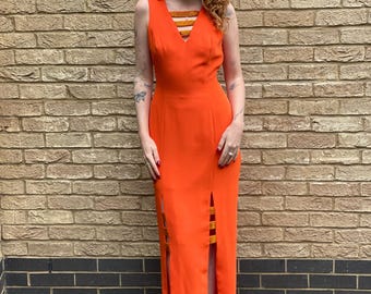 Könnte beinhalten: Ein langes orangefarbenes Maxikleid mit V-Ausschnitt und einem braunen Samtstreifen-Detail. Das Kleid hat einen Schlitz an der Seite und wird von einer Person mit roten Haaren getragen.