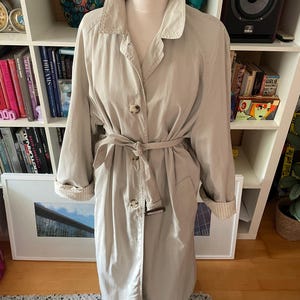 May include: A long, beige trench coat with a belt and a button fastening. The coat has a classic design and is made from a lightweight fabric.