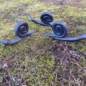 Może przedstawiać: Trzy metalowe ślimaki w ciemnoszarym kolorze są ułożone na łóżku z zielonego mchu. Ślimaki są wykonane w realistycznym stylu z detalicznymi muszlami i ciałami.