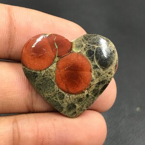 Puede incluir: Un cabujón de jaspe rojo y verde en forma de corazón con un patrón único de círculos rojos sobre un fondo verde y negro.