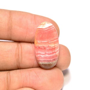 Puede incluir: Una gema ovalada con capas de tonos rosa, blanco y marrón. La superficie pulida revela un patrón de bandas, mostrando las variaciones naturales de color y textura. La gema se sostiene en una mano.