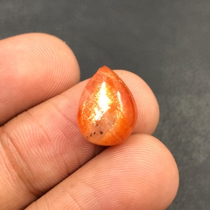 Może przedstawiać: Kabochoński szlif kamienia słonecznego w kształcie łzy, w kolorze jasno pomarańczowym i złotym. Kamień ma mieniący się, tęczowy efekt.