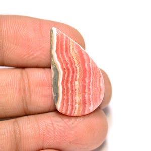 Puede incluir: Una piedra preciosa pulida en forma de lágrima, con un llamativo patrón de bandas rosas, blancas y beige. La piedra tiene una superficie lisa y brillante y un aspecto natural y orgánico. La piedra preciosa se sostiene en una mano.