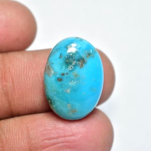Puede incluir: Gema ovalada de turquesa con superficie pulida. La piedra presenta un color azul brillante con inclusiones de matriz marrón y gris. La gema se sostiene entre los dedos sobre un fondo blanco.