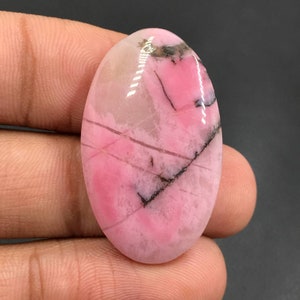 Puede incluir: Una piedra preciosa ovalada en cabujón de color rosa y blanco con vetas negras. La piedra preciosa tiene una superficie lisa y pulida.