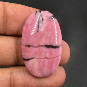 Puede incluir: Una piedra preciosa ovalada cabujón rosa y negra con acabado pulido. La piedra tiene un patrón de remolino de rosa y negro.